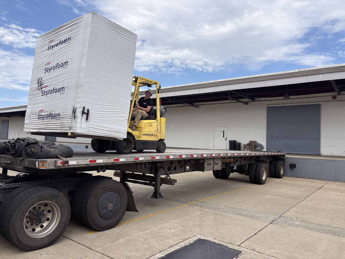 Yellow forklift loading large blocks of surplus Styrofoam onto a flatbed trailer at an industrial facility
